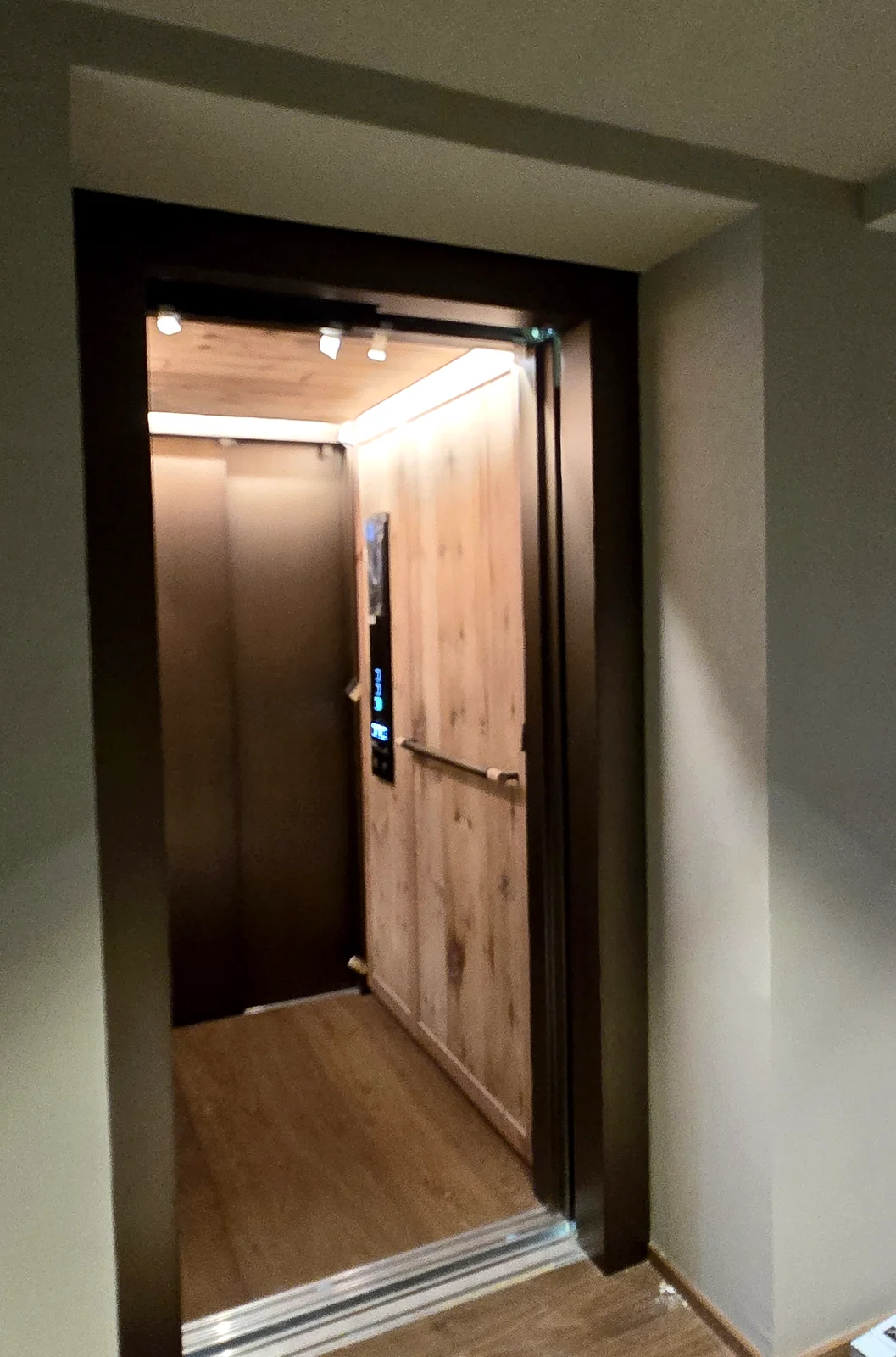 Open elevator portal with dark patinated brass frame and timber cabin lining at Golf Academy Grassau